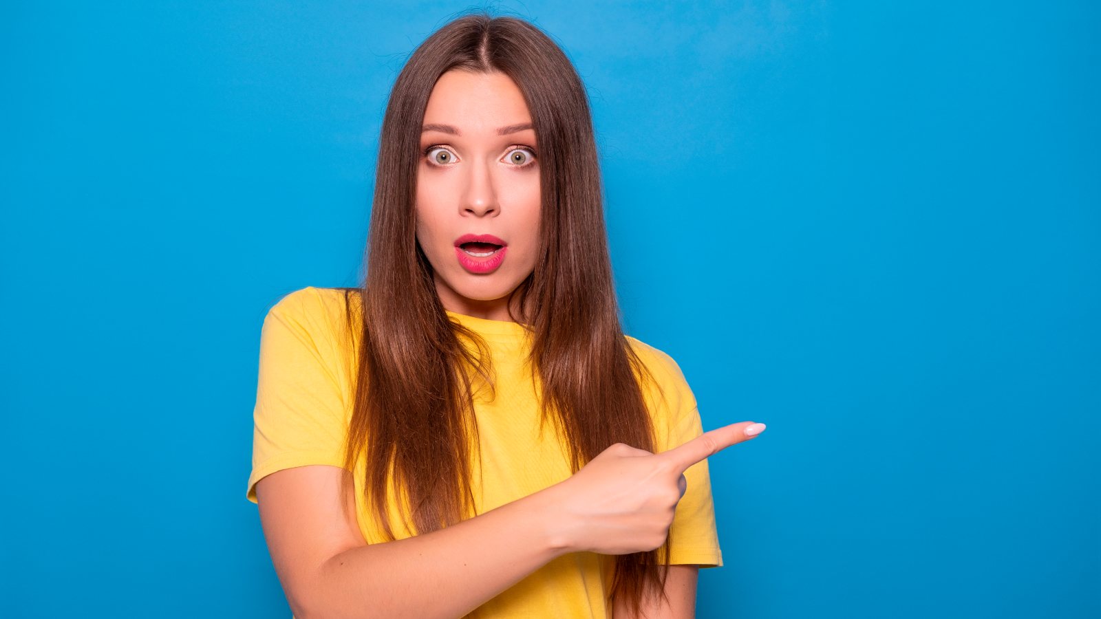 Cute brunette woman with long hair posing in yellow t-shirt on a blue background. Emotional portrait. She smiles happily with flawless white teeth and points finger on something